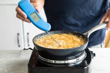Someone holding a thermometer in a brown, bubbling liquid in a small pan.