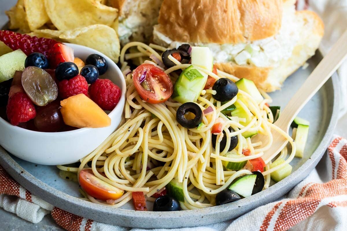 A plate with California pasta salad, fruit, chips and a croissant sandwich.