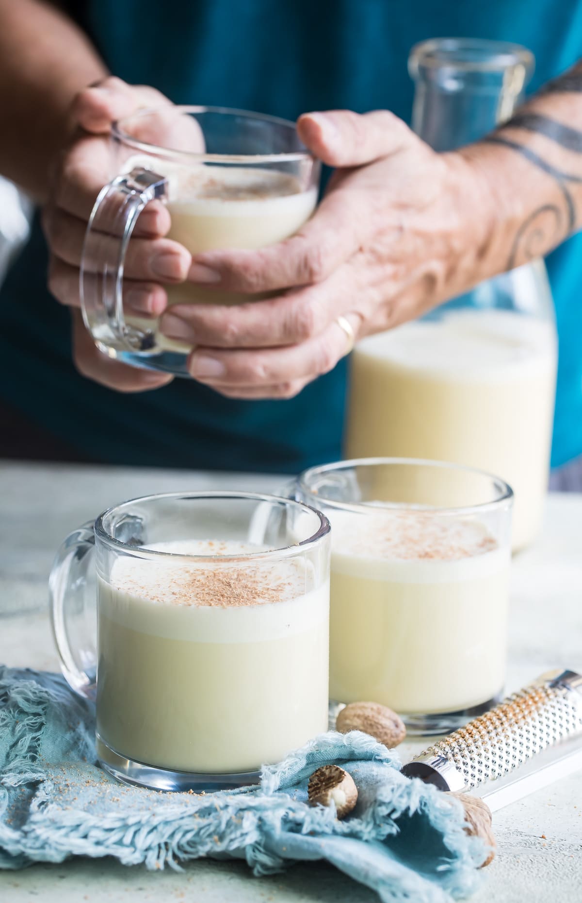 Someone holding a mug of eggnog with two cups of eggnog on a table.