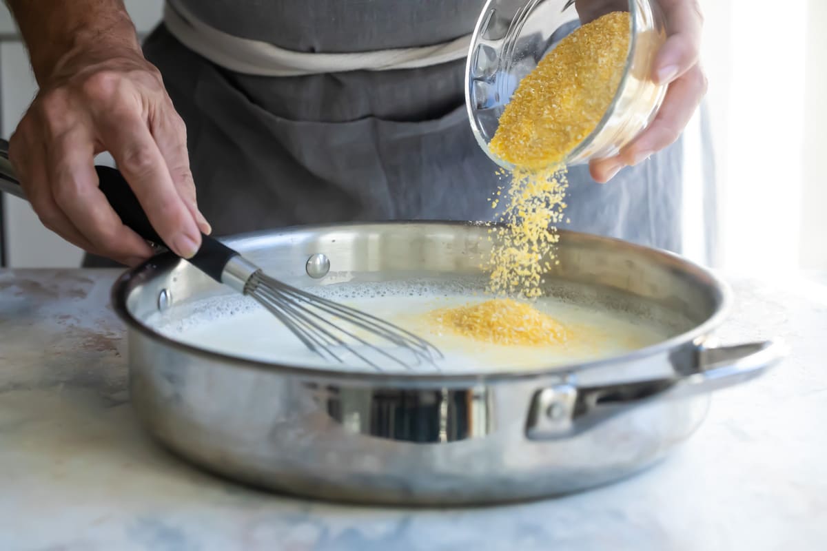 Grits being added to a silver pan with white liquid.