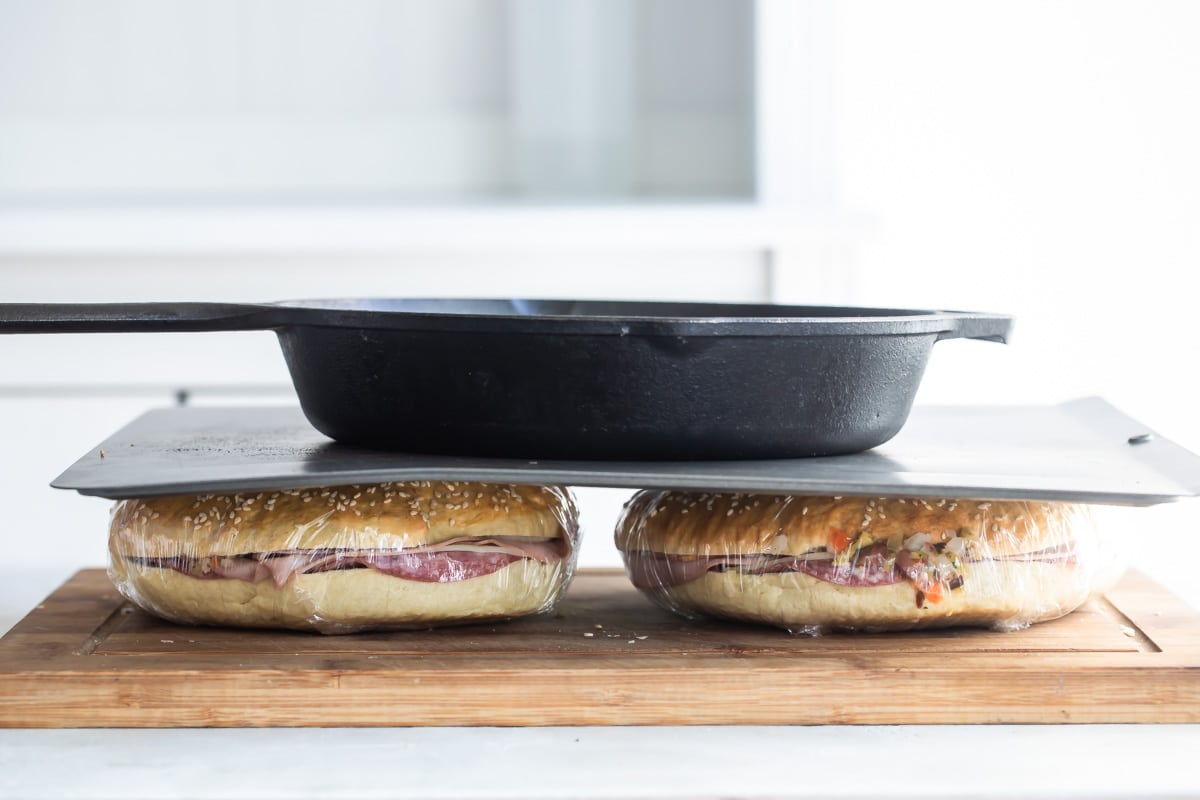 Muffaletta sandwiches being pressed down between a wooden cutting board and cast iron pan.