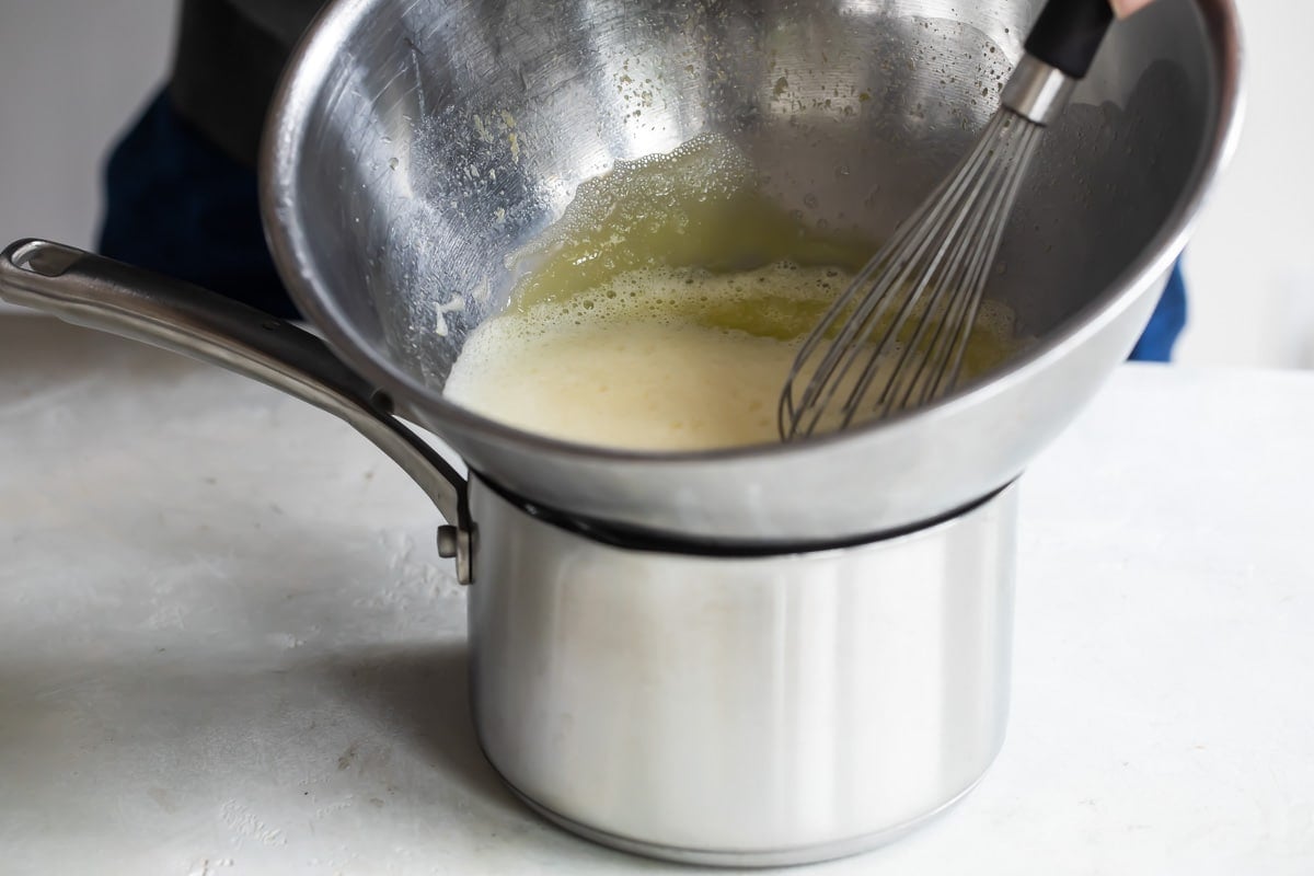 Yellow liquid being whisked in a bowl over a saucepan.