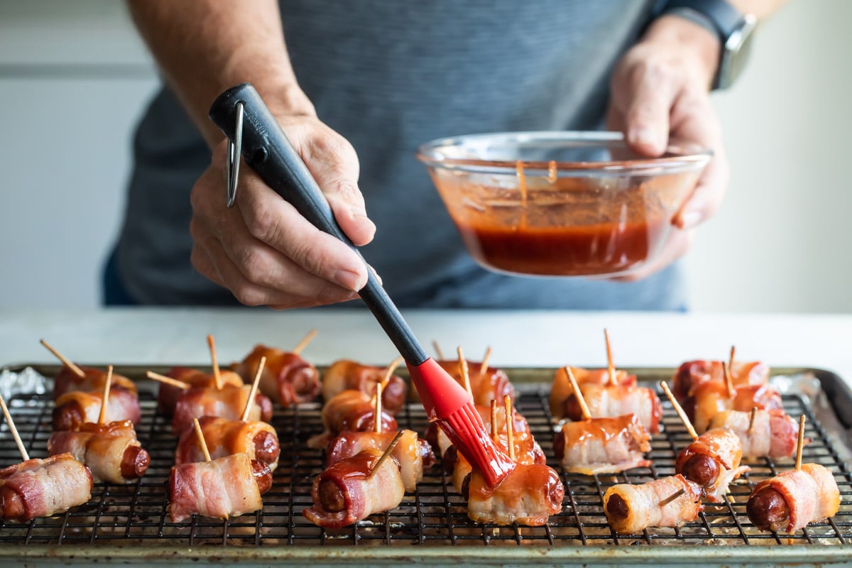 Someone putting sauce on bacon wrapped smokies on a cooling rack over a foil covered baking sheet.