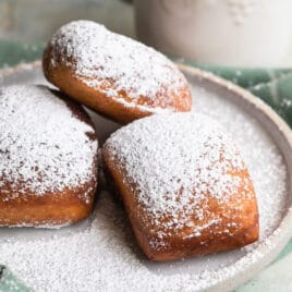 Beignets on a plate next to a coffee mug with liquid in it.