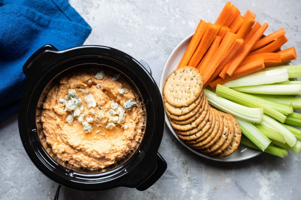 Cauliflower buffalo dip in a crockpot next to a bowl of carrots, celery and crackers.