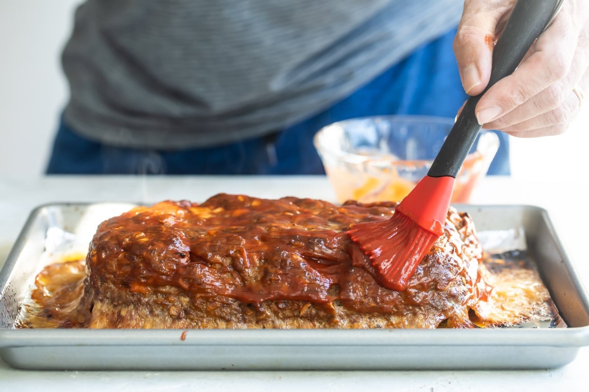 Someone brushing sauce on cheesy meatloaf.