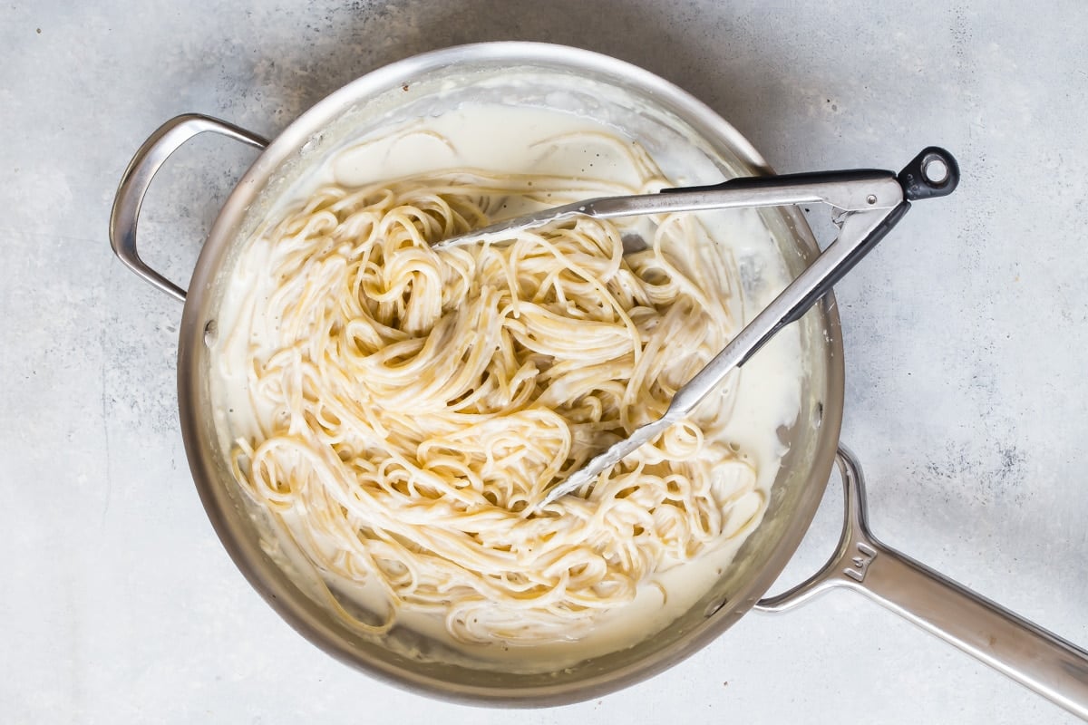Pasta with alfredo sauce in a silver skillet with tongs.