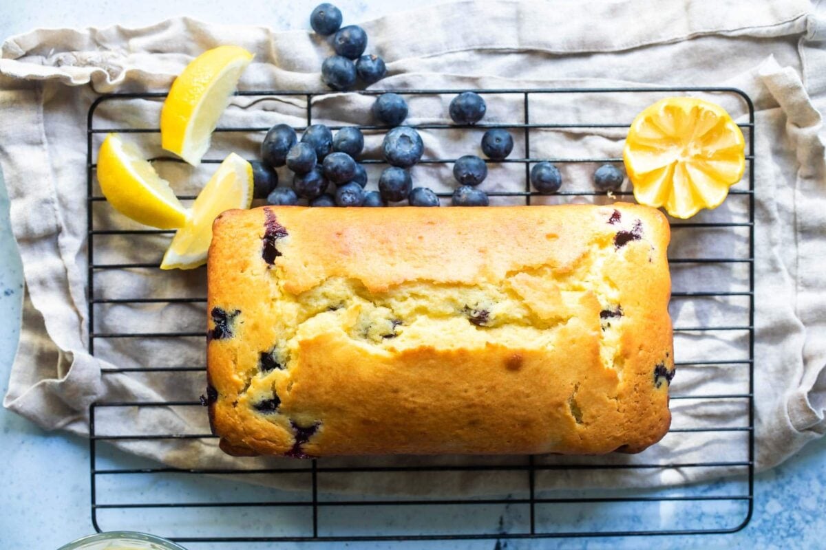 A loaf of blueberry lemon yogurt cake on a cooling rack surrounded by blueberries.
