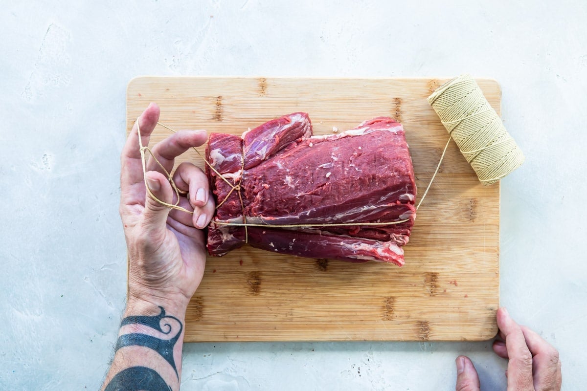 A beef tenderloin being tied on a wooden cutting board.