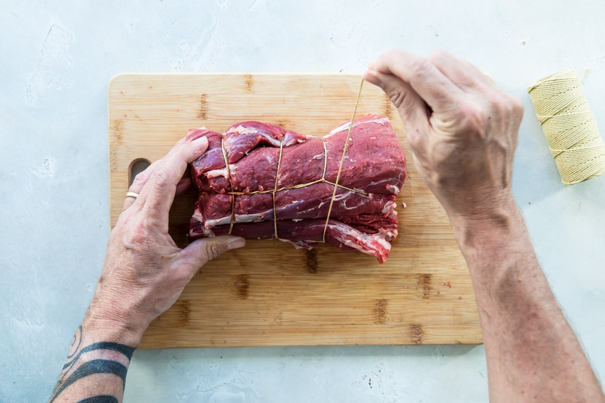 A beef tenderloin being tied on a wooden cutting board.