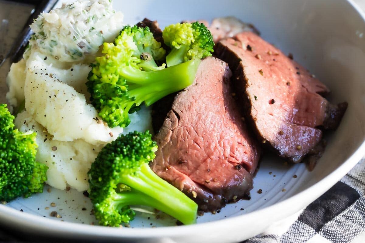 Slices of roast beef tenderloin on a plate with broccoli and mashed potatoes.