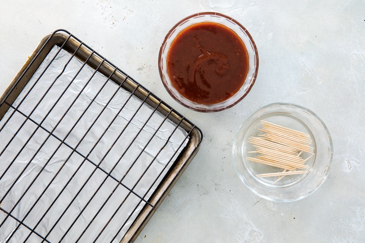 Barbecue sauce and toothpicks in small glass bowls next to a baking sheet with with a cooling rack on it.