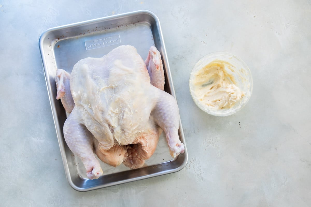 A whole chicken on a baking sheet with a bowl of garlic butter spread next to it.