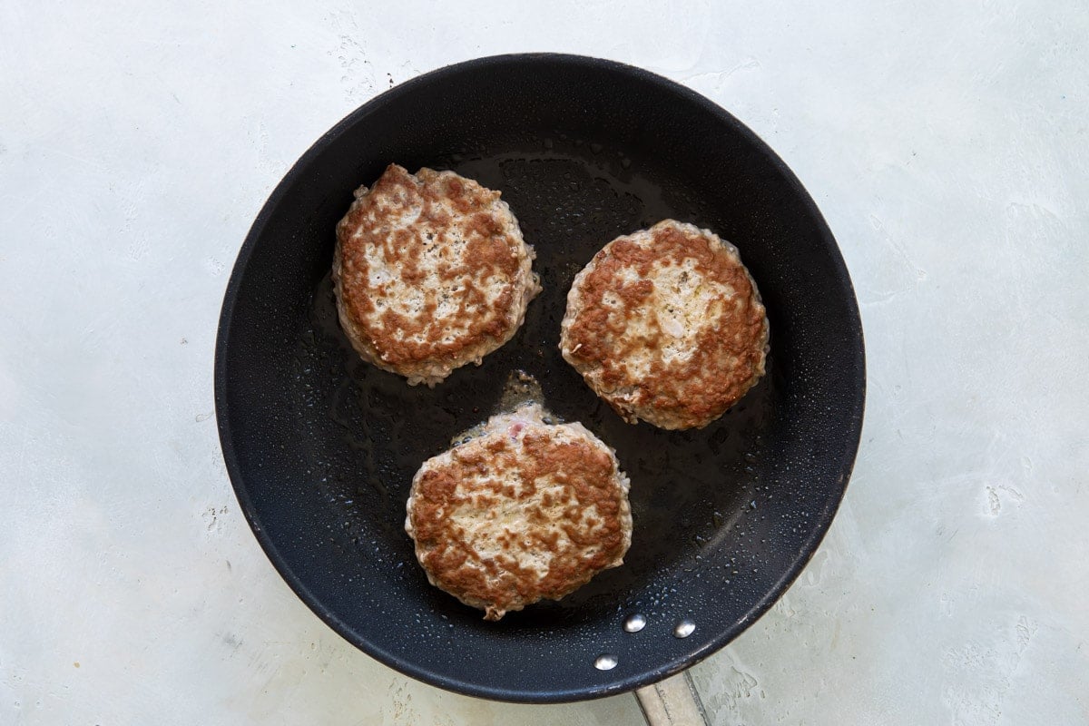 Three pork burgers being cooked in a black skillet.