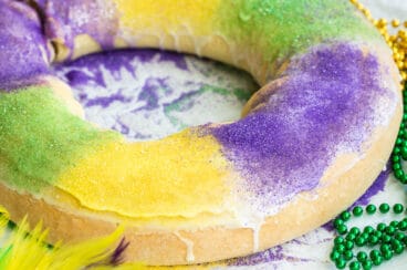 A decorated king cake on a white counter surrounded by Mardi Gras decorations.