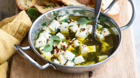 Marinated feta in a silver bowl on a wood board with slices of bread.