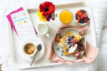 A Mother's Day breakfast tray with crepes, orange juice, coffee, fresh flowers, and a greeting card.