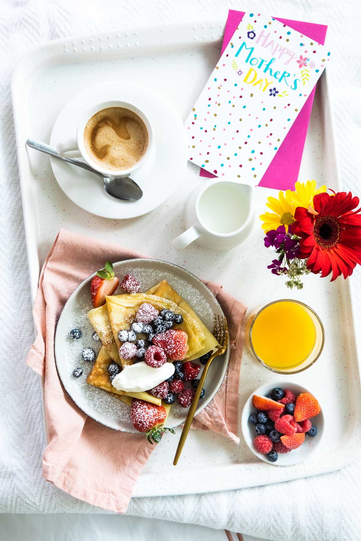 A Mother's Day breakfast tray with crepes, orange juice, coffee, fresh flowers, and a greeting card.