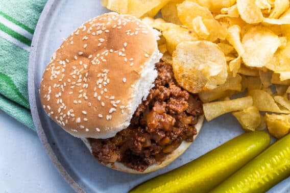 Plates of Sloppy Joes on plates with chips and pickles nearby.
