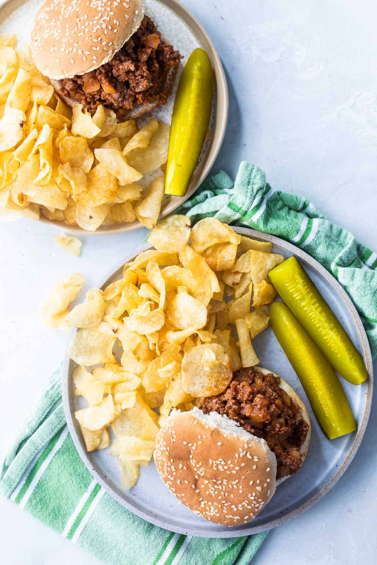 Plates of Sloppy Joes on plates with chips and pickles nearby.