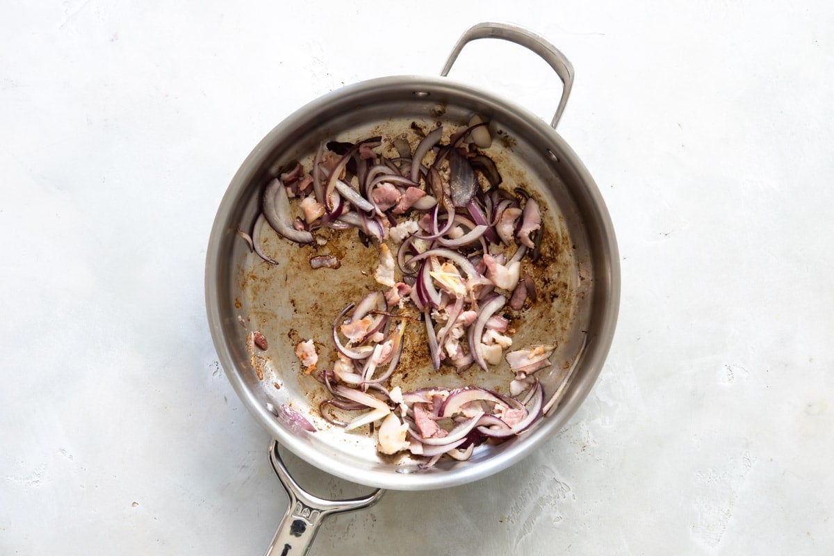 Onion, garlic and bacon being cooked in a silver skillet.