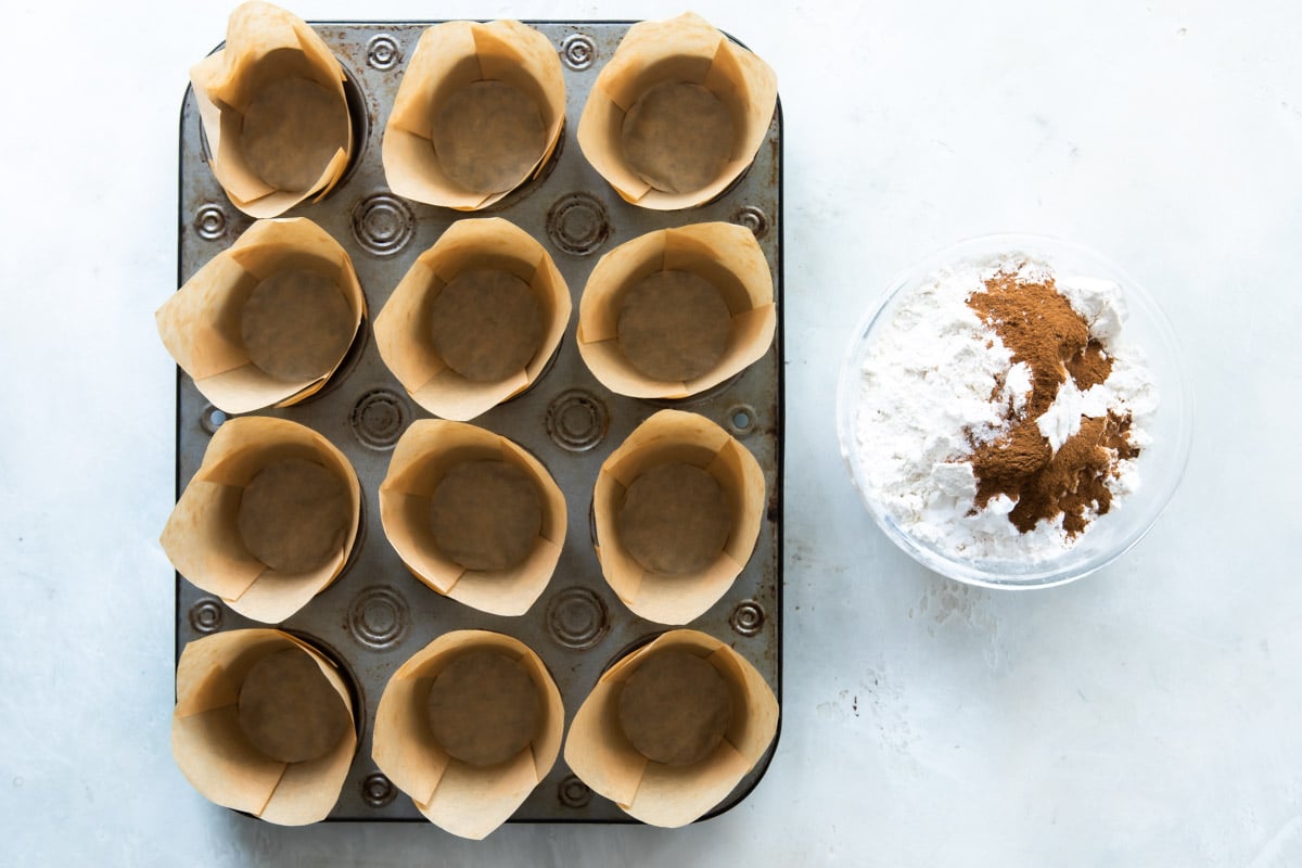 A muffin tin with paper cups in it next to a bowl of dry ingredients.