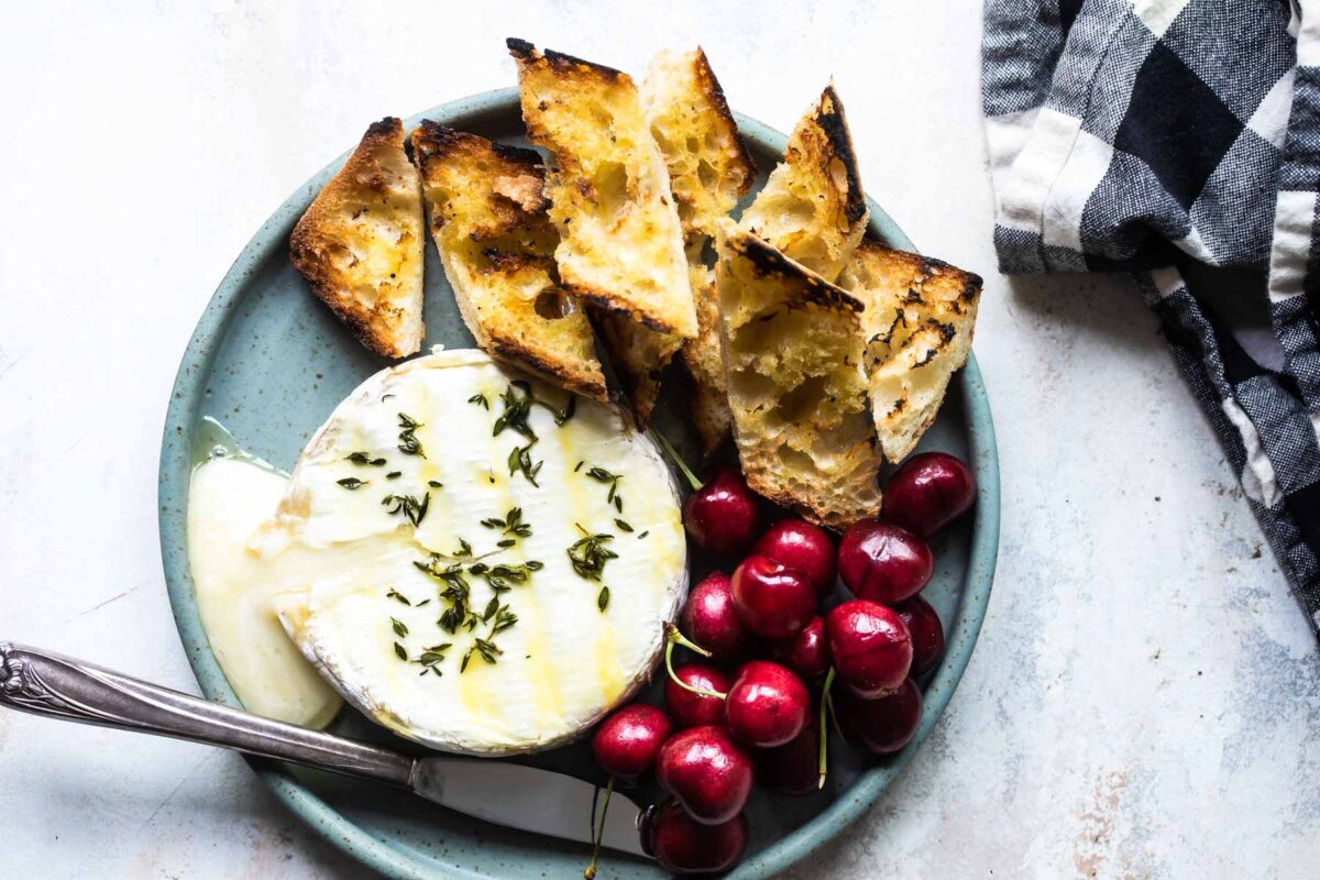 Baked brie on a blue plate with cherries and toasted bread pieces.