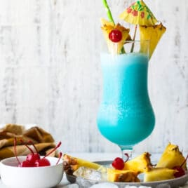 A Blue Hawaiian cocktail surrounded by pineapple and maraschino cherries.
