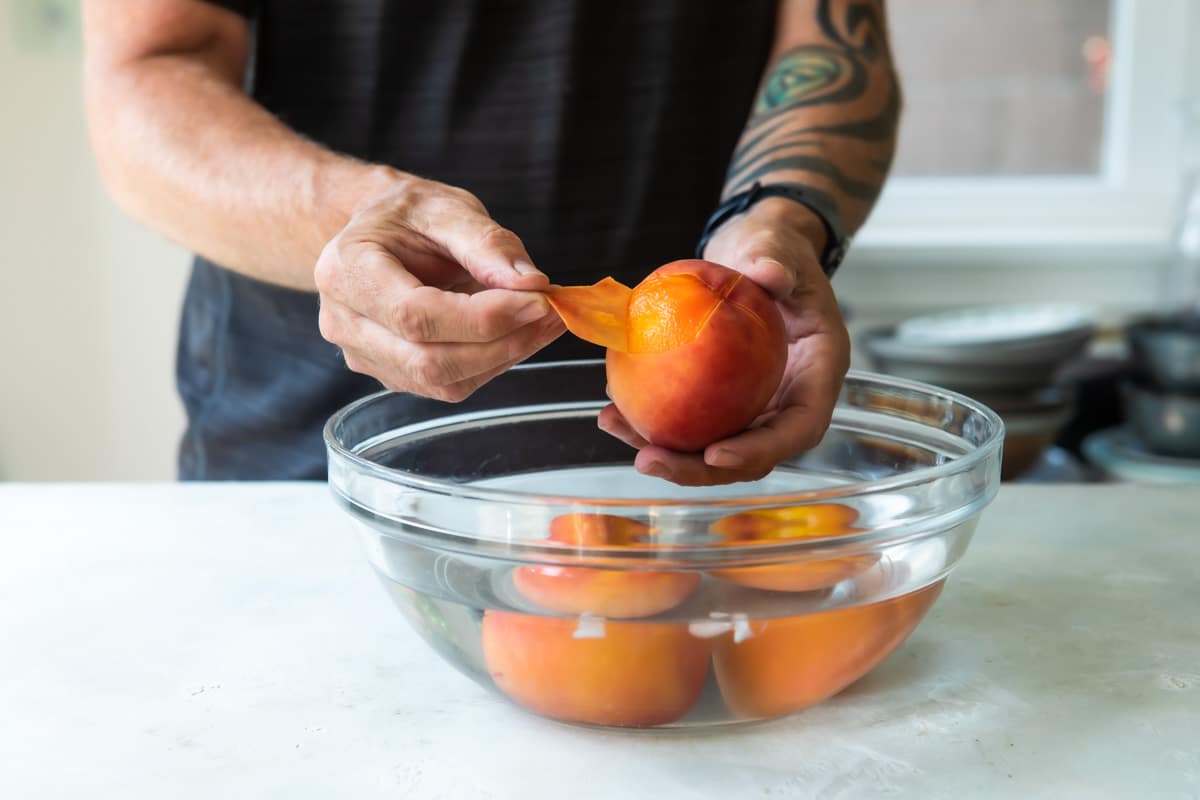 Peaches being peeled over water in a clear bowl.