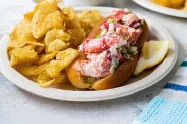 White plates with lobster rolls, potato chips, and lemon slices piled on top.