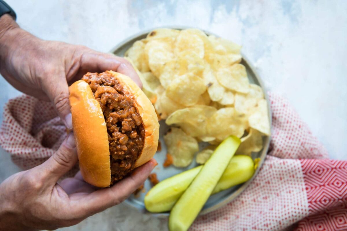 Someone holding a sloppy joe over a plate with pickles and chips.