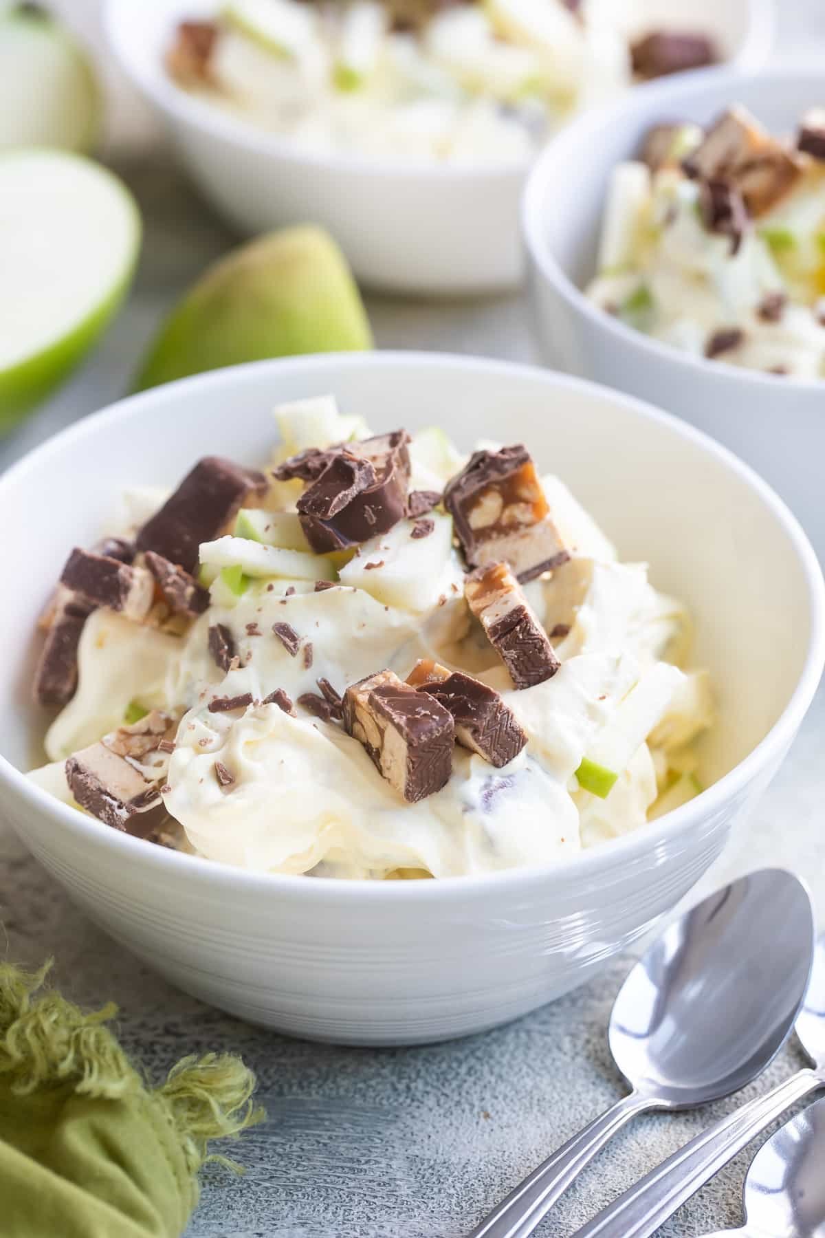Snickers salad in three bowls surrounded by apple pieces.
