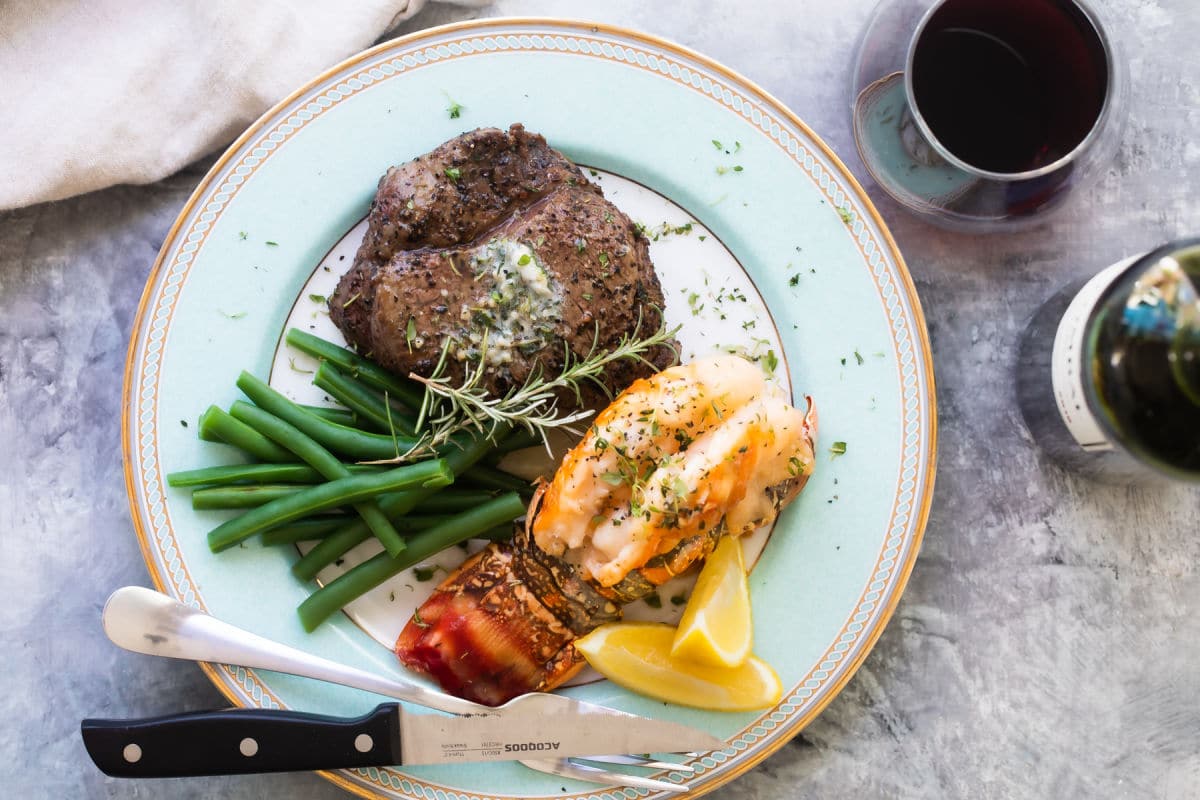 Surf and Turf (a filet mignon with a lobster tail) on a white plate with green beans with wine in the background.