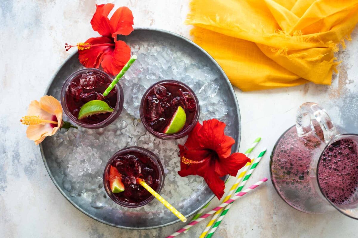 Glasses of Agua de Jamaica on a tray with hibiscus flowers and straws.