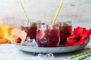 Glasses of Agua de Jamaica on a tray with hibiscus flowers and straws.