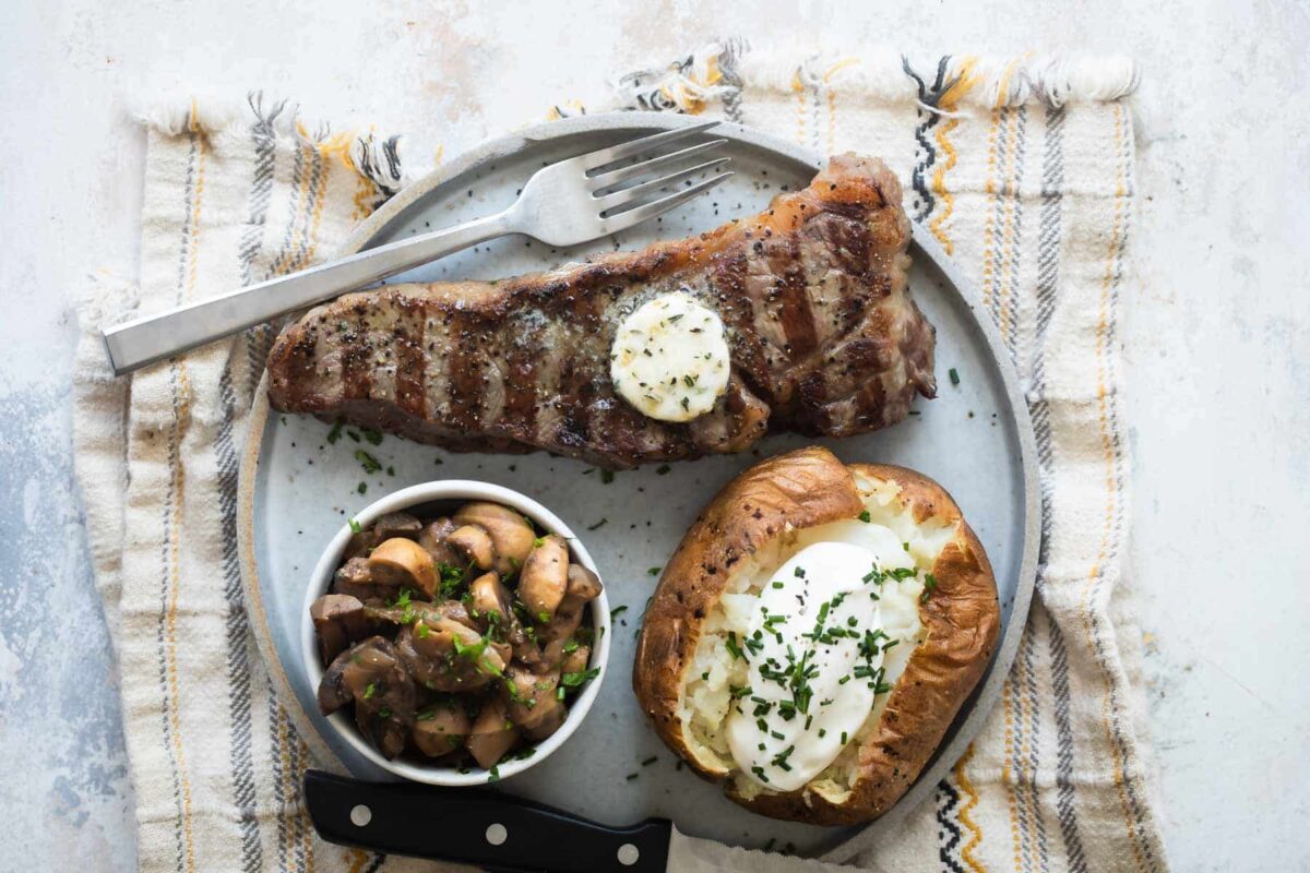 A New York Strip Steak, baked potato, and mushrooms on a plate.