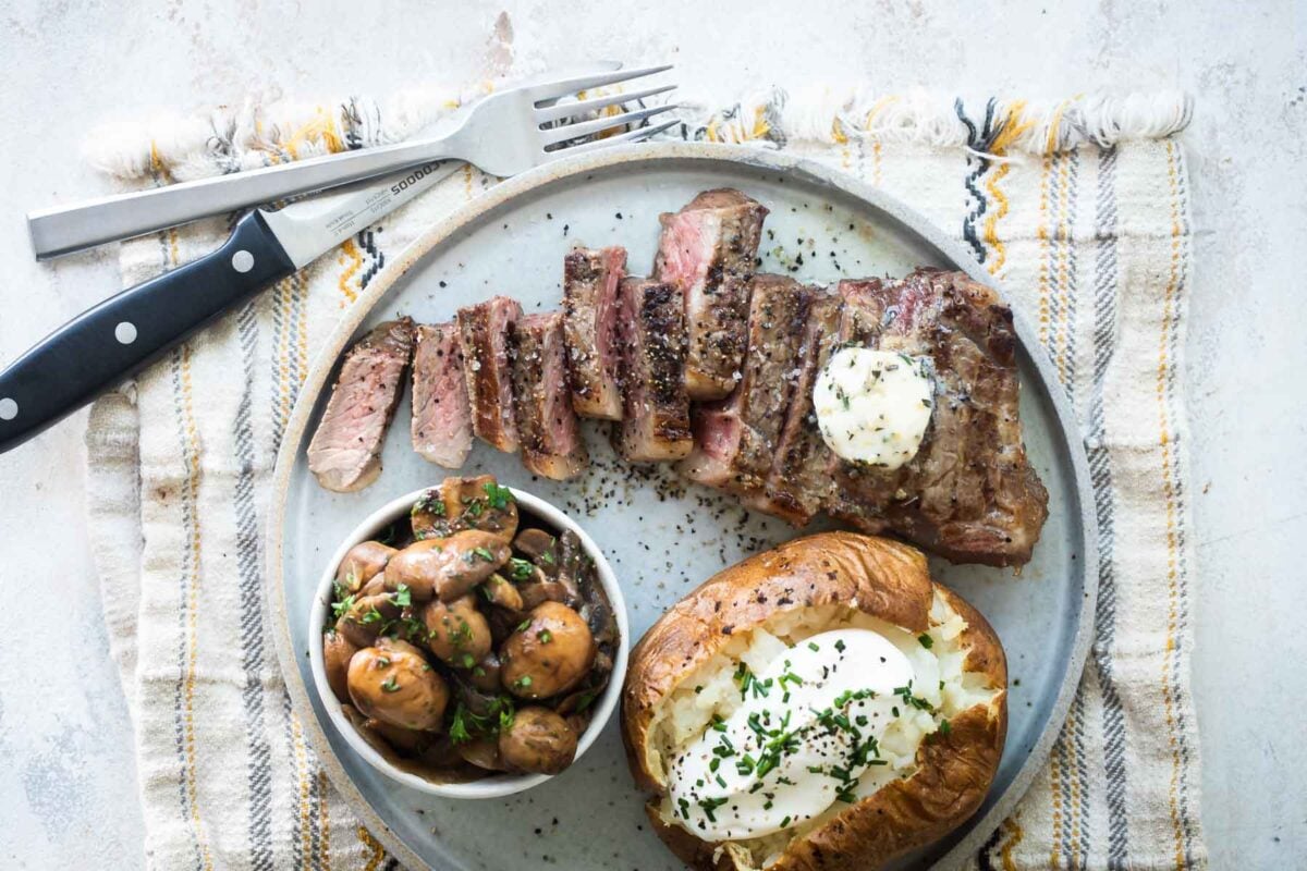 A plate with New York strip steak, a baked potato, and mushrooms.
