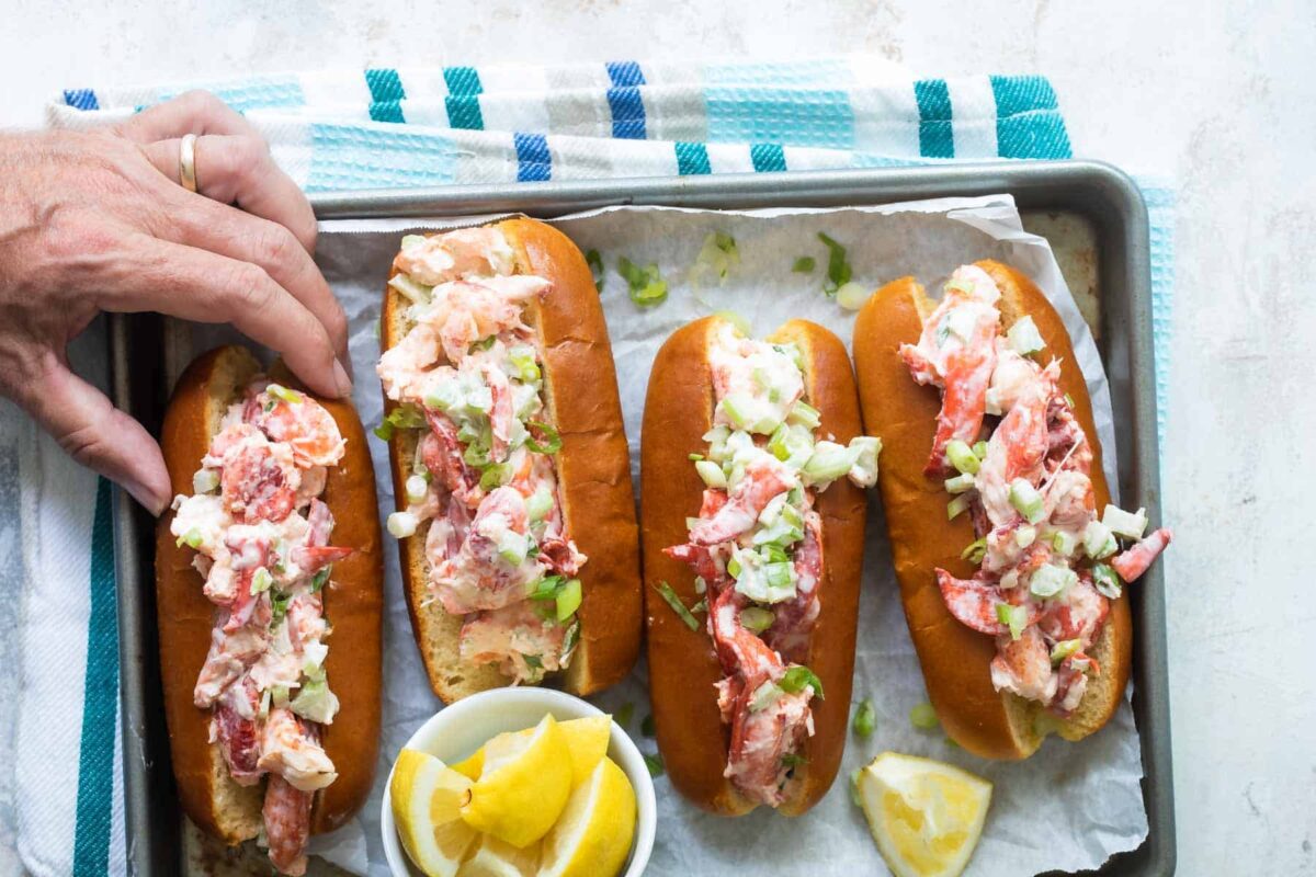 A tray of lobster rolls with a bowl of lemons and a hand grabbing one roll.
