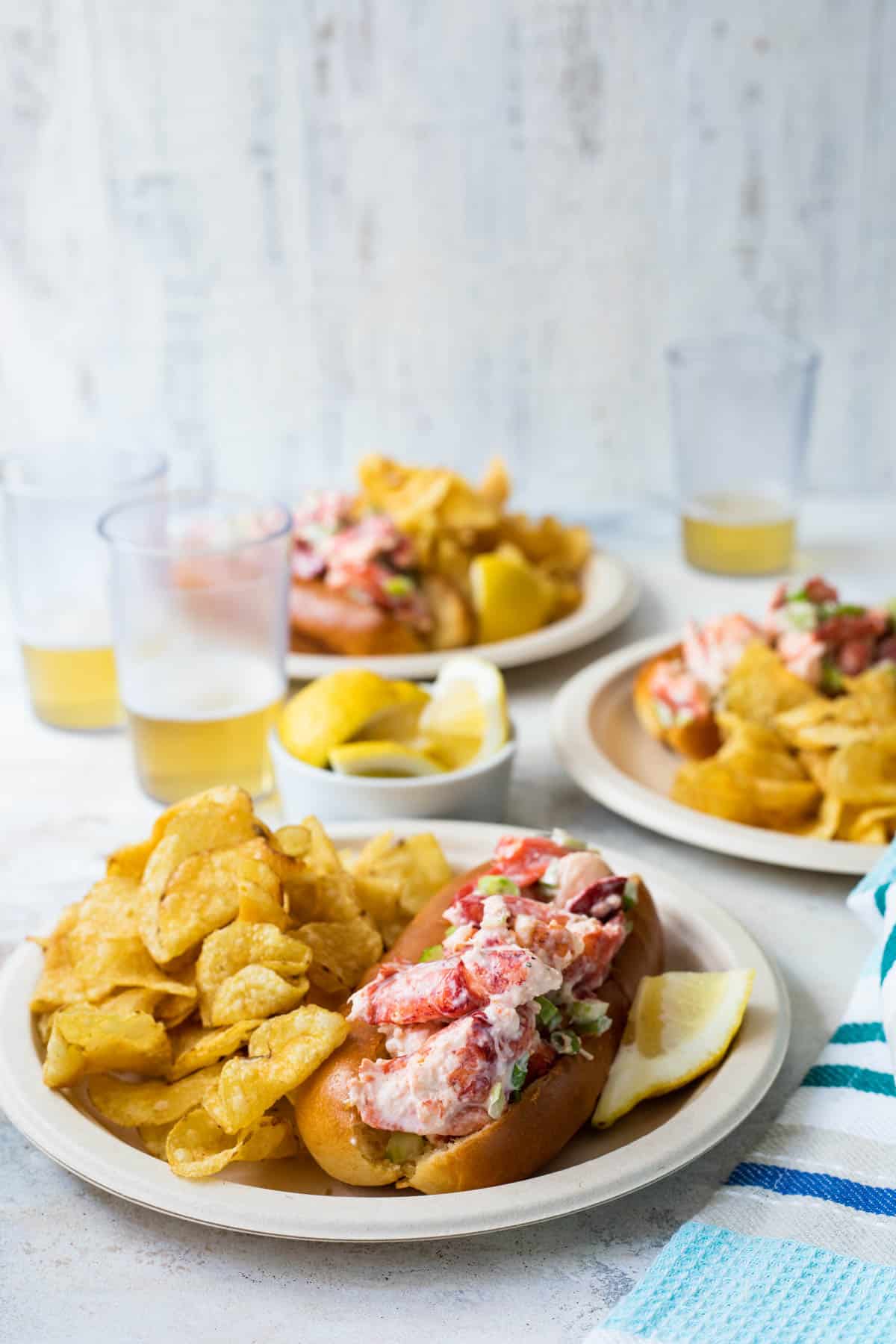 White plates with lobster rolls, potato chips, and lemon slices piled on top.