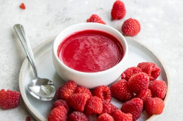 Raspberry coulis in a white dish on a white plate with raspberries.