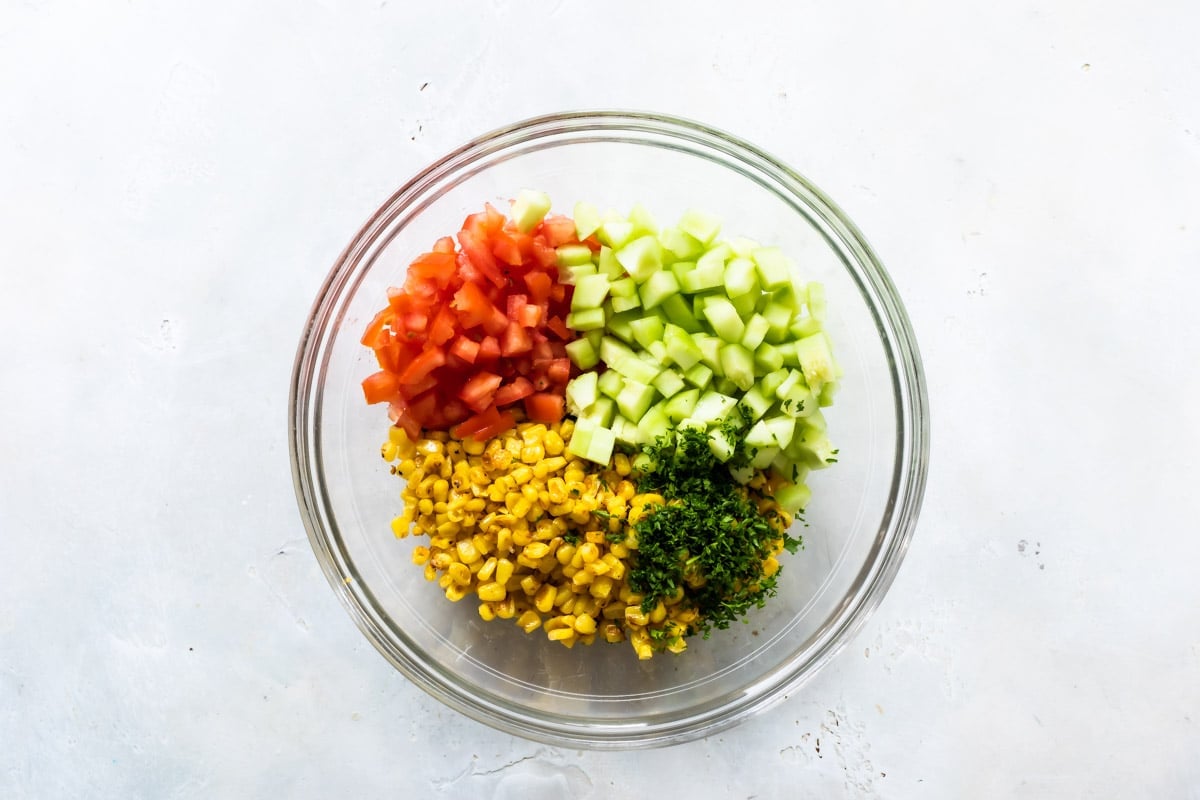 A clear glass bowl full of corn, tomatoes, cucumbers, and parsley.
