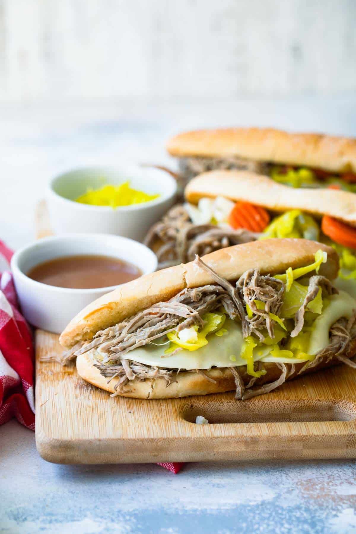 A cutting board with several Italian Beef sandwiches and a bowl of au jus nearby.