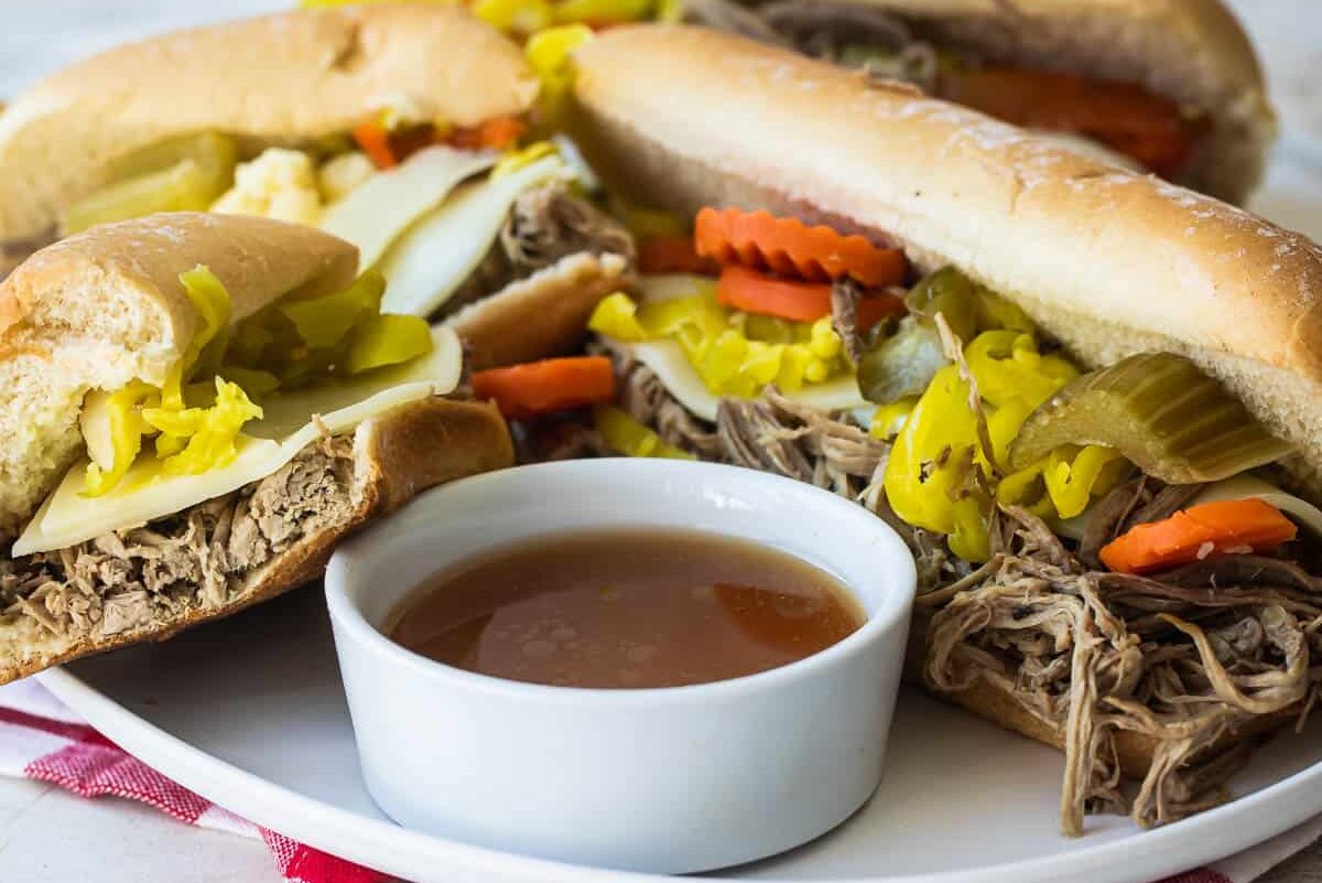 A platter with several Italian Beef sandwiches and a bowl of au jus nearby.