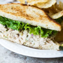A tuna salad sandwich on a plate next to chips and a pickle spear.