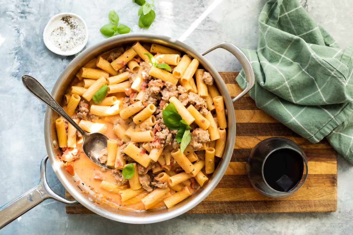 A large skillet filled with Rigatoni with Sausage with a green napkin and wine next to it.