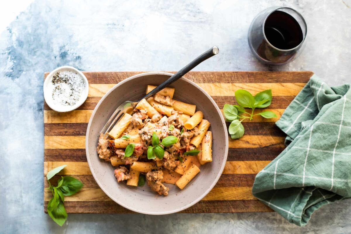 A bowl filled with Rigatoni with Sausage with a green napkin and wine next to it.