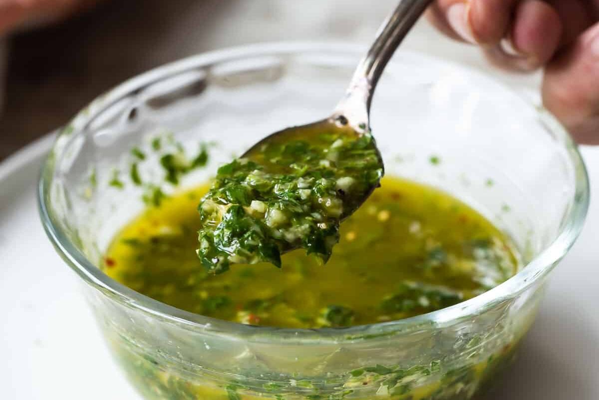 Chimichurri sauce being spooned out of a small clear glass bowl.