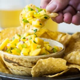 Someone scooping mango salsa out of a bowl with a chip.