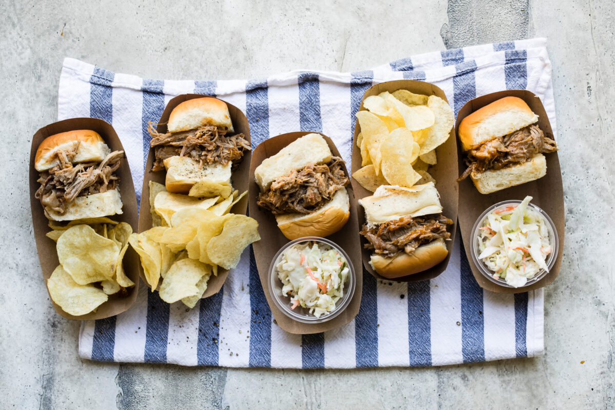 Slow cooker pulled pork sandwiches with chips and coleslaw in brown trays.