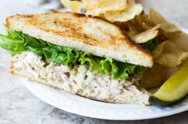 A tuna salad sandwich on a plate with a pickle spear and potato chips.